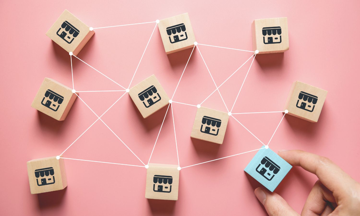 Hand placing a block with a shop icon among connected blocks on a pink background, symbolizing a real estate franchise network.