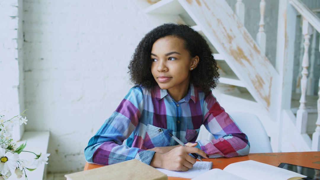 A person sits at a bright, sunlit table with papers, looking pensive as they weigh the pros and cons of their home selling strategy.